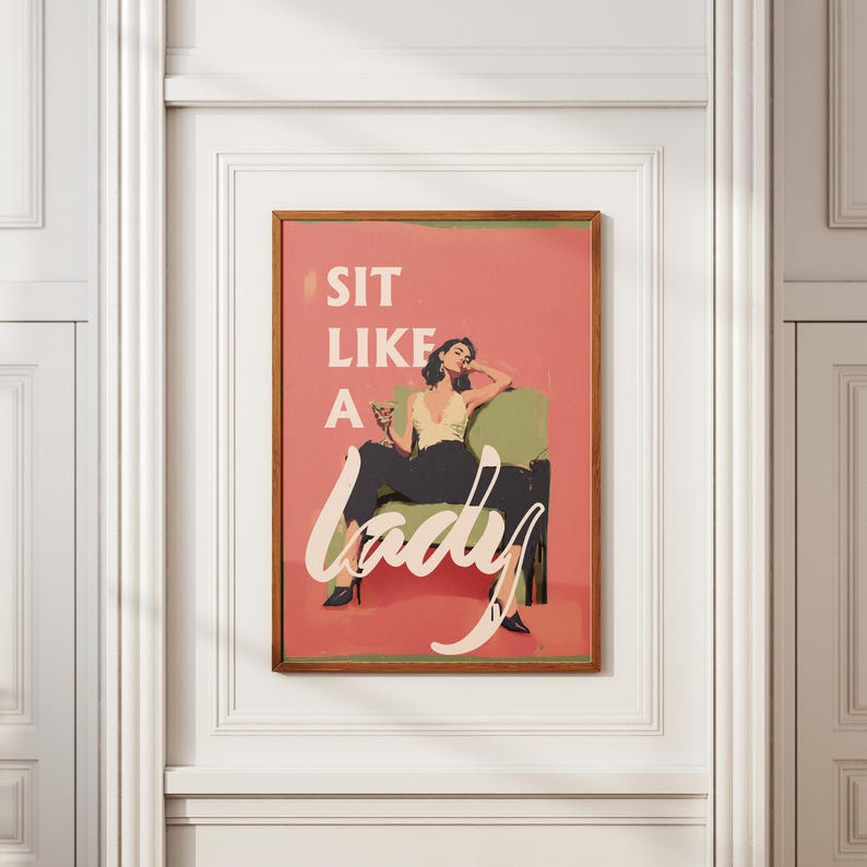 May include: A framed art print with the text "SIT LIKE A lady" in white script. The artwork depicts a woman in a yellow top and black pants, seated in a green chair, holding a drink. The background is a salmon pink.