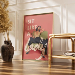 May include: A framed art print with a pink background and the text "SIT LIKE A LADY." The artwork features a woman in a chair, holding a drink. The frame is gold, and the print is displayed in a room with a vase and a small table.