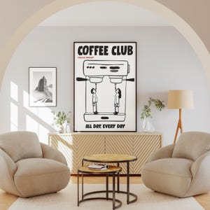 Op de afbeelding: Een zwart-wit ingelijste print met de tekst "COFFEE CLUB" en "ALL DAY, EVERY DAY". De print toont een koffiemachine met twee mensen eronder. De kamer omvat twee beige fauteuils, een houten kast en twee ronde salontafels.