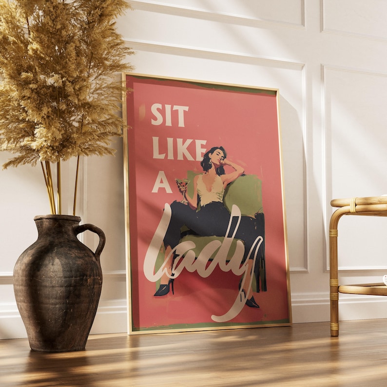May include: Framed art print with a retro design. The print features a woman in a chair with the words "SIT LIKE A LADY" in white script on a pink background. The frame is gold, and the artwork is displayed in a room with a vase and a side table.
