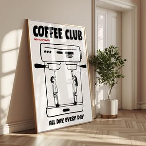 Op de afbeelding: Een ingelijste poster met een gouden rand, leunend tegen een muur. De poster toont een zwart-wit illustratie van een koffiemachine met twee figuren die naar de tuiten kijken. De woorden "COFFEE CLUB" en "ALL DAY, EVERY DAY" staan op de poster.