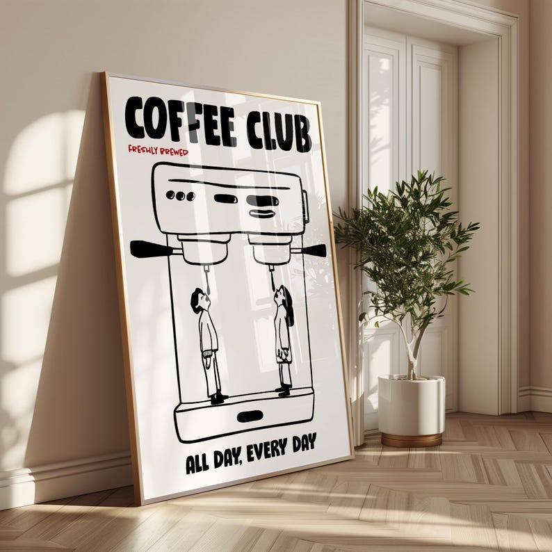 Op de afbeelding: Een ingelijste poster met een gouden rand, leunend tegen een muur. De poster toont een zwart-wit illustratie van een koffiemachine met twee figuren die naar de tuiten kijken. De woorden "COFFEE CLUB" en "ALL DAY, EVERY DAY" staan op de poster.