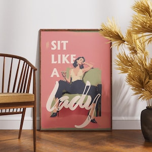 May include: A framed art print with a pink background and the words "SIT LIKE A LADY" in white script. The artwork depicts a woman in a cream top and dark pants, seated on a green chair, holding a cocktail. The frame is brown.
