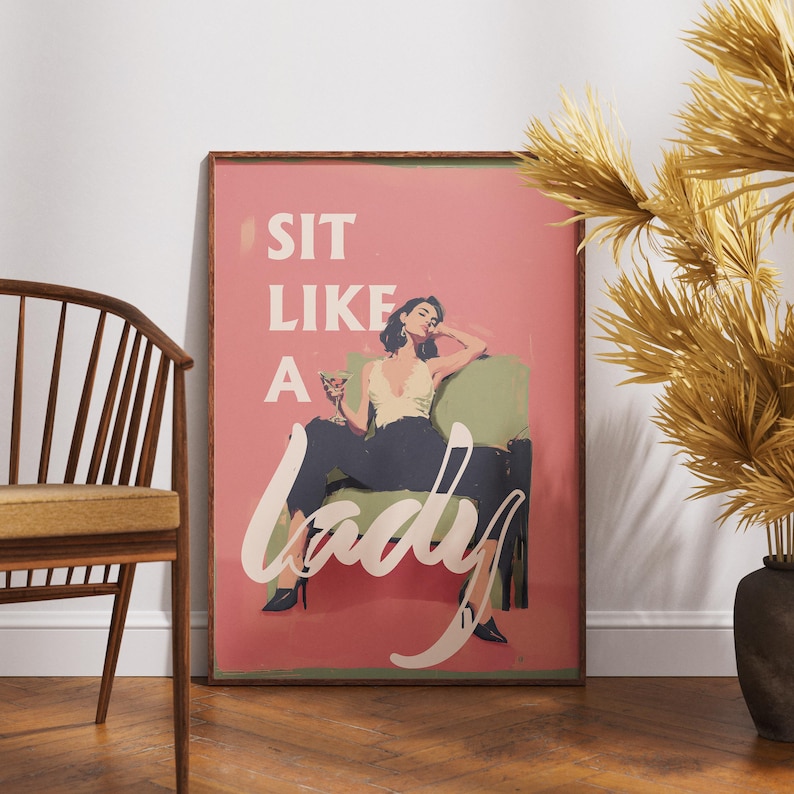 May include: A framed art print with a pink background and the words "SIT LIKE A LADY" in white script. The artwork depicts a woman in a cream top and dark pants, seated on a green chair, holding a cocktail. The frame is brown.