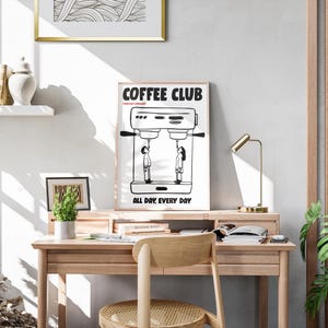 Op de afbeelding: Een zwart-wit print met de tekst "COFFEE CLUB" en "ALL DAY. EVERY DAY." De print toont een koffiemachine met twee figuren eronder. De print staat op een houten bureau met een kleine plant en een gouden bureaulamp.