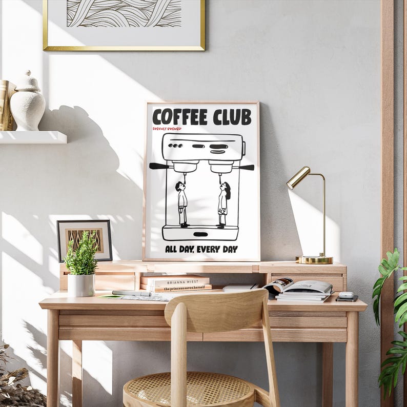 Op de afbeelding: Een zwart-wit print met de tekst "COFFEE CLUB" en "ALL DAY. EVERY DAY." De print toont een koffiemachine met twee figuren eronder. De print staat op een houten bureau met een kleine plant en een gouden bureaulamp.