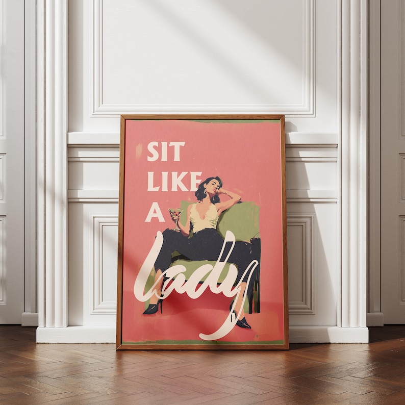 May include: A framed art print with a retro design. The print features a woman in a yellow top and black pants, seated in a green chair, with the words "SIT LIKE A LADY" in white script on a pink background.