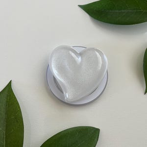 May include: A white heart-shaped phone grip featuring a smooth, glossy finish with a subtle shimmering, pearlescent texture. The heart is mounted on a circular, collapsible base, providing a functional accessory for mobile devices.