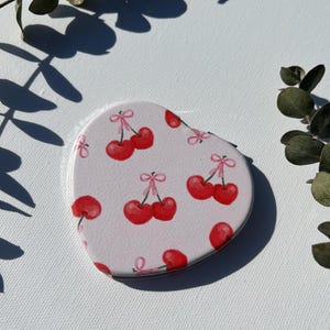 May include: A heart-shaped compact mirror with a light pink background and a pattern of red cherries with pink bows. The mirror is surrounded by green leaves and their shadows.