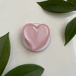 May include: A heart-shaped phone grip featuring a smooth, glossy, light pink finish with a subtle shimmering effect. The heart is mounted on a circular white base, providing a functional accessory for mobile devices with a clean and simple design.