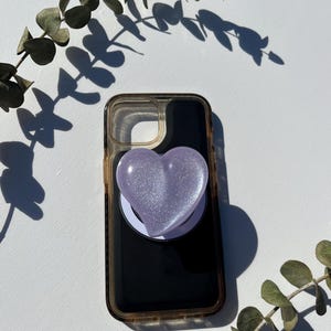 May include: A clear phone case with a black interior and a light purple heart-shaped phone grip. The heart grip has a glittery, iridescent finish. The phone case is on a white surface with green foliage.