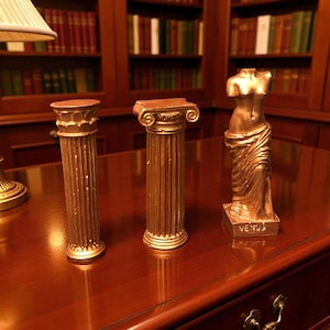 Könnte beinhalten: Drei goldfarbene Dekorationsartikel auf einer Holzoberfläche: zwei Miniatur-Säulen und eine Venus-Statue. Die Säulen weisen detaillierte Schnitzereien auf. Die Statue zeigt einen drapierten weiblichen Torso. Das Wort "VENUS" steht auf dem Sockel.