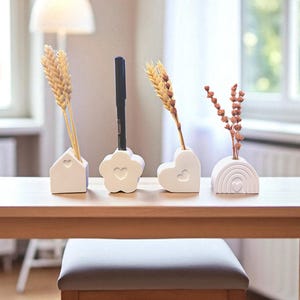 May include: Four white ceramic decorative holders in the shape of a house, flower, heart, and rainbow, each with a heart cutout. The holders contain wheat stalks, a black pen, and dried flowers. The items are arranged on a wooden table.