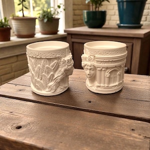 Puede incluir: Dos candelabros cilíndricos de color blanquecino con diseños de relieve clásicos. Uno presenta el rostro de una mujer con follaje, el otro una fachada de edificio clásico. Ambos tienen un borde de clave griega en la parte superior y están sobre una mesa de madera.