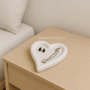 May include: A white heart-shaped jewelry tray with a pair of teardrop earrings and a silver necklace with a single clear gemstone. The earrings have a dark blue stone. The tray is on a light wood surface.