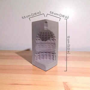 May include: A gray concrete sculpture of a building with a domed roof. The sculpture is 5.12 inches tall and 2.16 inches wide. The building's architectural details are visible. The sculpture is on a wooden surface.