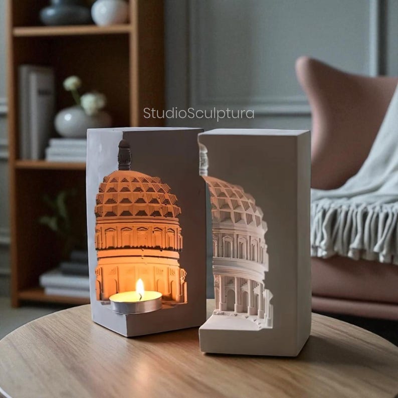 May include: Two gray, rectangular candle holders with a Capitol building design. One holder has a lit candle inside, illuminating the building's details. The other holder shows the building's exterior. The text "StudioSculptura" is visible.