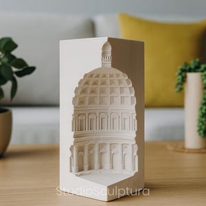 May include: A white architectural sculpture of a domed building, possibly a capitol building, with detailed relief work. The sculpture is on a wooden table, with the text "StudioSculptura" visible. The background includes a couch and plants.