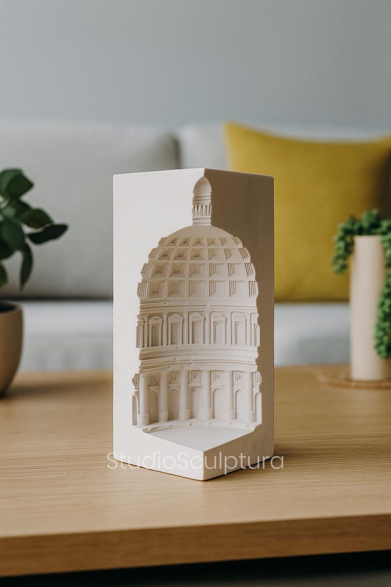 May include: A white architectural sculpture of a domed building, possibly a capitol building, with detailed relief work. The sculpture is on a wooden table, with the text "StudioSculptura" visible. The background includes a couch and plants.