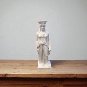 May include: A white statue of a woman in classical attire, standing on a rectangular base. The statue has a detailed headpiece and draped clothing. The sculpture is set against a light gray background and rests on a wooden surface.