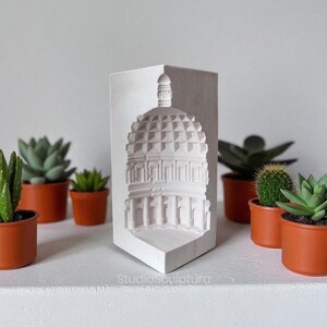 May include: A white, architectural relief sculpture of a domed building, likely a historical landmark, with detailed columns and a textured roof. The sculpture is displayed on a white shelf, surrounded by small potted succulents in terracotta pots. The text "StudioSculptura" is visible.