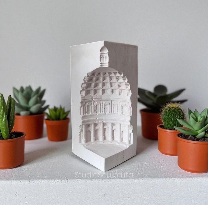 May include: A white, architectural relief sculpture of a domed building, likely a historical landmark, with detailed columns and a textured roof. The sculpture is displayed on a white shelf, surrounded by small potted succulents in terracotta pots. The text "StudioSculptura" is visible.