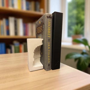 May include: A white bookend with a detailed architectural design supports two books. One book is titled "The Hobbit" by J.R.R. Tolkien, and the other is a black hardcover. The books are on a light-colored wooden surface, with a blurred bookshelf in the background.
