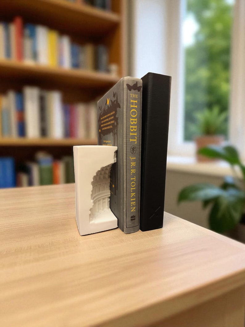 May include: A white bookend with a detailed architectural design supports two books. One book is titled "The Hobbit" by J.R.R. Tolkien, and the other is a black hardcover. The books are on a light-colored wooden surface, with a blurred bookshelf in the background.