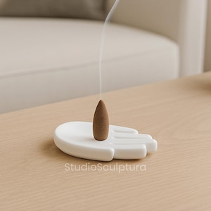 May include: A white ceramic hand-shaped incense holder with a brown incense cone emitting a wisp of smoke. The text "StudioSculptura" is printed on the holder. The holder is on a light brown wooden surface.