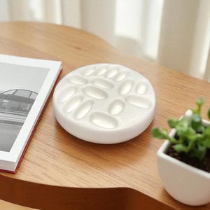 Op de afbeelding: Een wit keramisch verfpalet met meerdere ovale putjes ligt op een houten tafel. Een boek met een rode rug en een kleine potplant staan ook op de tafel. Het palet is ontworpen voor het mengen van verven.