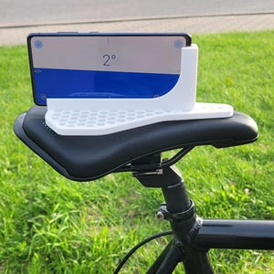 Op de afbeelding: Een witte telefoonhouder met een honingraatpatroon is gemonteerd op een zwart fietszadel. Een smartphone toont een blauw-wit scherm met het nummer "2°" zichtbaar. De fiets is zwart met zilveren onderdelen, tegen een achtergrond van groen gras en een weg.