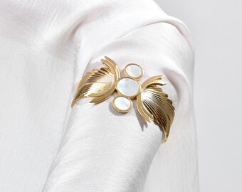 May include: A gold-coloured metal brooch with a leaf and circular design, featuring white accents, is pinned to white fabric. The brooch is a decorative accessory, adding a touch of elegance to the garment.