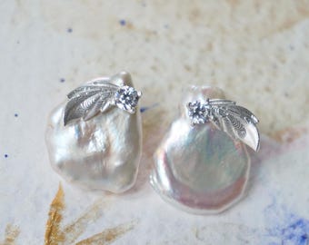 May include: Two large, irregular-shaped white pearl earrings. Each earring features a small, clear gemstone and a silver-colored, leaf-shaped accent. The earrings are displayed on a textured, off-white surface with gold and blue accents.