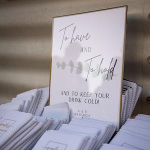 May include: A sign with the text "To have and to hold and to keep your drink cold!" with a sprig of eucalyptus and the date 04.13.2024. A basket filled with white can coolers with the text "A & D 04.13.2024" printed on them.