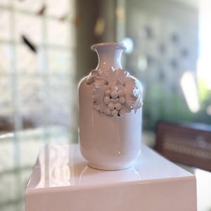 May include: A white ceramic vase with a grape cluster design. The vase is rectangular with a narrow neck and is displayed on a white platform. The grape cluster is raised and adds texture to the vase.