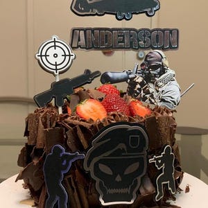 Op de afbeelding: Een chocoladetaart versierd met een militair thema. De taart is gegarneerd met verse aardbeien, een helikopter, een doel, een geweer en een soldatenfiguur. De naam "ANDERSON" staat op de taart.