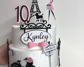 May include: Two-tiered white cake decorated with a black silhouette of the Eiffel Tower, a woman walking a poodle, a bicycle, and a sign that says "Paris". The cake also has a pink bow, a black number 10, and the name "Kynley" written on it.