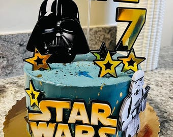 May include: A blue and gold Star Wars themed cake with a Darth Vader topper, a gold number 7, and the words "STAR WARS" on the front. The cake is decorated with gold stars and splatters of gold paint.