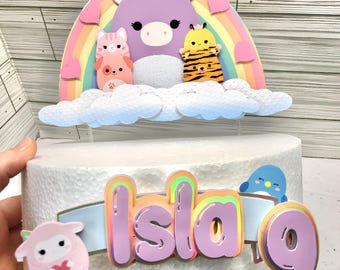 May include: A purple unicorn with a rainbow behind it and a cloud underneath. There are four other animal figures on the cloud, including a cat, a dog, a sheep, and a tiger. The word "Isla" is written in pink with a rainbow outline, and the number "9" is written in pink with a rainbow outline. The figures are on a white cake.