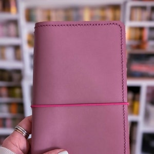 May include: A mauve leather journal with a pink elastic band closure. The journal has a stitched edge and a smooth, matte finish.