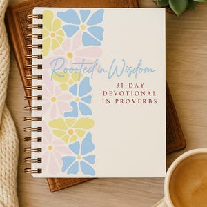 Puede incluir: Un cuaderno con espiral con un diseño floral en colores pastel, con el texto "Rooted in Wisdom" y "31-Day Devotional in Proverbs". El cuaderno descansa sobre un libro encuadernado en cuero, con una suculenta y una taza de café cerca.