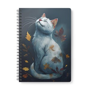 Puede incluir: Cuaderno de espiral con una ilustración de un gato blanco con los ojos cerrados, rodeado de hojas de otoño cayendo en tonos dorados, rojos y marrones. El gato es el foco central, sobre un fondo oscuro.