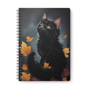 Pode incluir: Caderno espiral com uma ilustração de um gato preto com olhos amarelos olhando para cima. A imagem inclui folhas de outono caindo em tons de amarelo e vermelho. O fundo é cinza escuro e o caderno tem uma borda branca.
