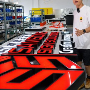 May include: A workshop scene featuring illuminated LED signs in various stages of completion. The signs are primarily red and black, with some white. The signs spell out words and phrases. A person stands next to the signs.