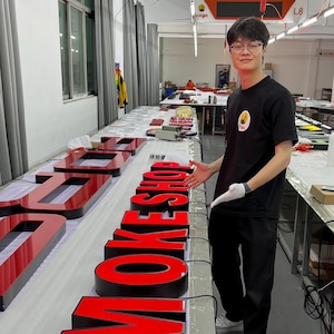 May include: Large, illuminated red and black letters spelling "SMOKE" are displayed on a table in a workshop. The letters are three-dimensional and appear to be made of acrylic or a similar material. A person is standing next to the letters.