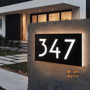 Könnte beinhalten: Moderne beleuchtete Hausnummerntafel mit der Nummer 347 in Weiß auf schwarzem Hintergrund. Die Tafel ist auf einem strukturierten Steinsockel montiert, wobei ein warmes Licht von hinter den Ziffern ausgeht. Die Hausnummer wird in einer modernen Schriftart angezeigt.