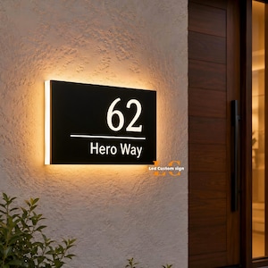 May include: A black illuminated address sign with the number 62 and the text "Hero Way" in white. The sign is rectangular and mounted on a textured wall, with a warm light emanating from behind it. The sign is near a wooden door.
