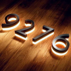 May include: Black metal illuminated house numbers 9276, with a warm white glow. The numbers are displayed on a wooden surface, creating a modern and elegant aesthetic. The numbers are approximately 6 inches tall.