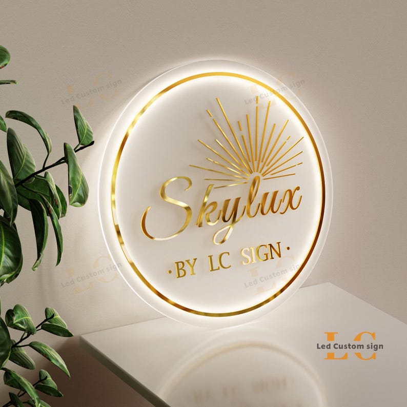 May include: A circular, illuminated sign with a white face and gold detailing. The sign displays the word "Skylux" in gold script, with "BY LC SIGN" underneath. A gold sunburst design is above the word "Skylux". The sign is lit with a soft glow.