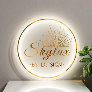 May include: A round, illuminated sign with a gold border and the name "Skylux" in elegant script. The sign also features the text "BY LC SIGN" and a gold sunburst design. The sign is mounted on a white surface.